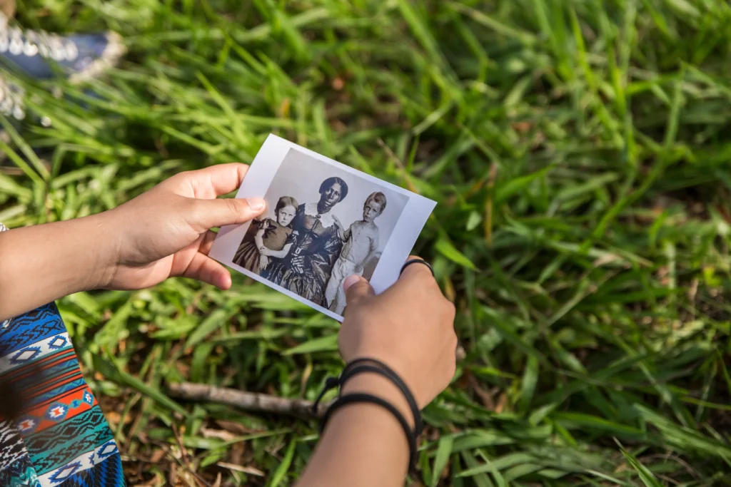 Close de duas mãos segurando uma pequena fotografia antiga em preto e branco sobre um fundo de grama verde. A foto na mão retrata três pessoas. A pessoa que segura a foto usa pulseiras pretas e uma roupa com estampa geométrica azul e vermelha.