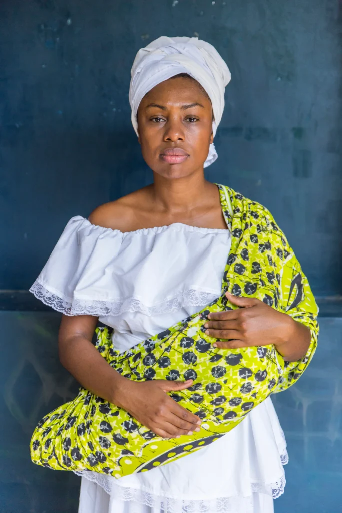 A artista Renata Felinto em performance para a 11ª MITsp. Ela aparece de perfil, com o rosto e ombros cobertos por argila ou barro claro, sobre um fundo branco.